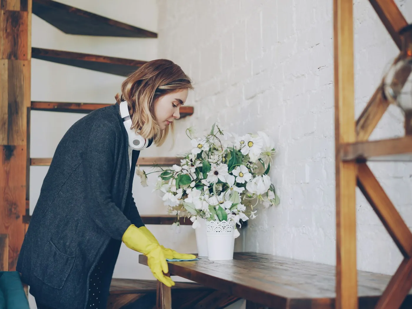 A cleaner in yellow rubber gloves wiping a wooden table beside a vase of white flowers
